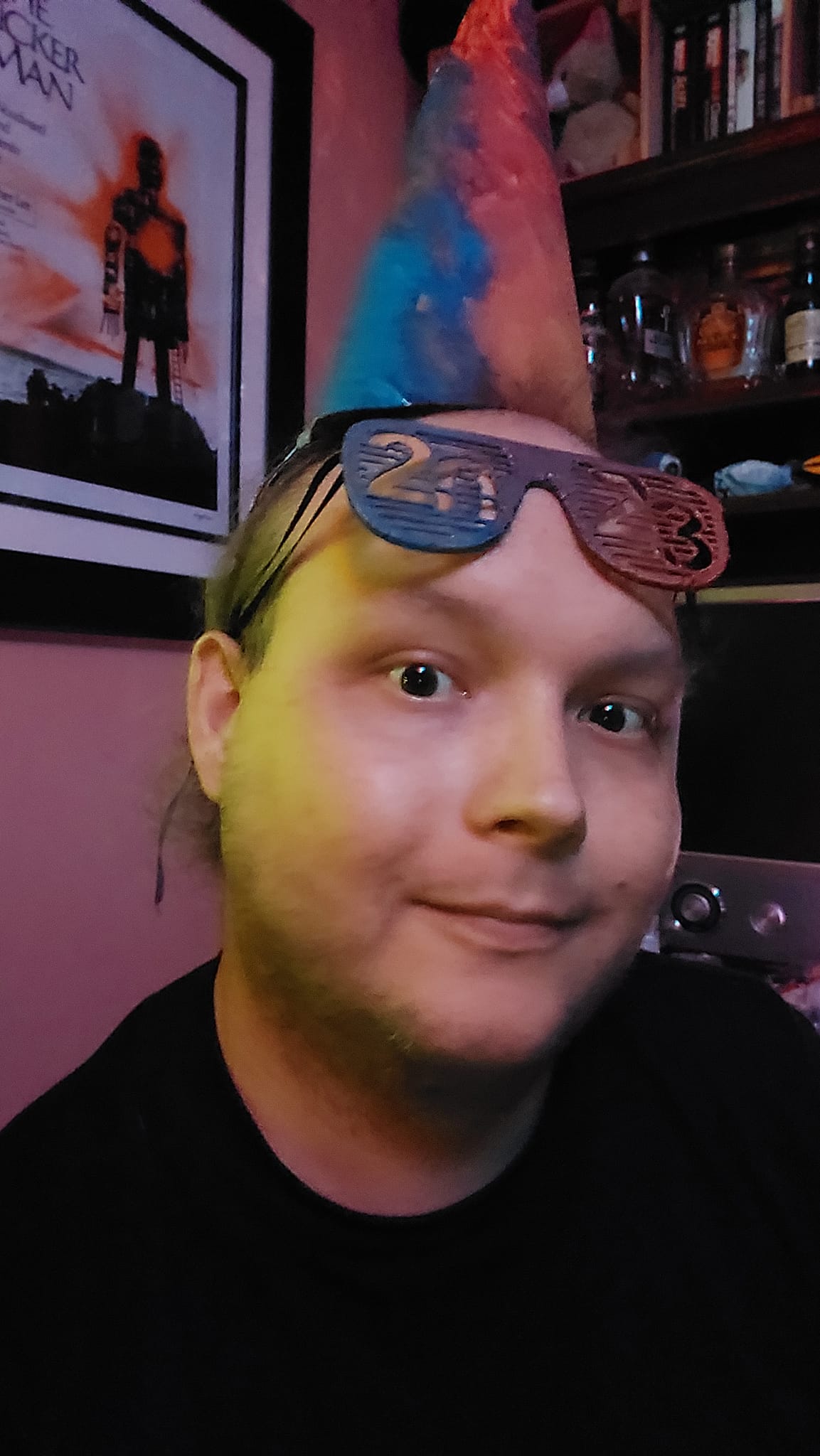 A man sits in a corner, wearing both the hat and painted glasses, the glasses are raised to his forehead the lighting is purple